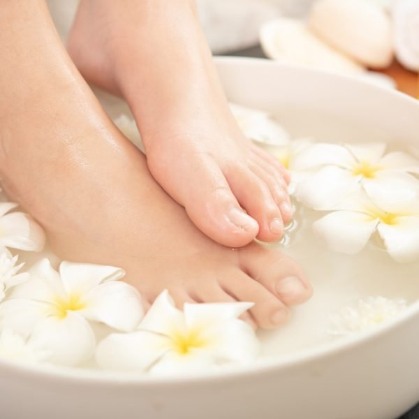 closeup view of woman soaking her feet in dish with water and flowers on wooden floor. Spa treatment and product for female feet and hand spa. white flowers in ceramic bowl.